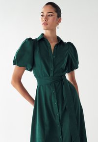 Zelena kratka oblekica s kratkimi rokavi, z gumbom spredaj, z vezanim pasom v pasu in teksturirano tkanino, ki vključuje subtilen lesk ter napihnjene rokave.