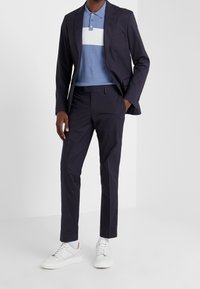 Marinblazer över en ljusblå polo med vita ränder, parat med mörka byxor och vita sneakers. Mjukt tyg, skräddarsydd passform.