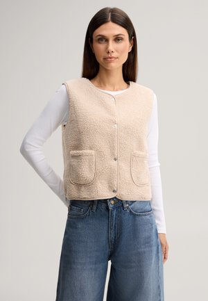 Donna indossa un gilet in pile beige con bottoni e tasche anteriori sopra una camicia bianca a maniche lunghe e jeans blu larghi su sfondo semplice.