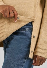 Bež pletena pulover z rebrastimi manšetami in bočnimi gumbi, ki ima mehko teksturo. V kombinaciji z modrimi jeans hlačami v oprijetem slogu.
