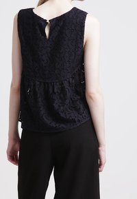 Navy blue lace top with a round neckline, keyhole back, and flared hem; features a layered texture and intricate floral pattern.