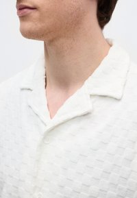 Κοντινό πλάνο του λαιμού και του σαγονιού ενός άντρα που φοράει υφασμάτινο λευκό πουκάμισο με γιακά και κουμπιά, με φόντο μια απλή επιφάνεια.