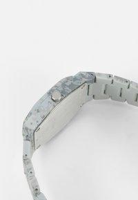 Reloj de patrón gris con una caja rectangular de plástico, parte trasera de metal plateado y una correa flexible gris con segmentos enlazados.
