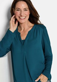 Türkisfarbige Bluse mit langen Ärmeln und V-Ausschnitt, mit Raffung an den Schultern, aus einem glatten Stoff gefertigt. Minimalistisches Design, lässige Passform.