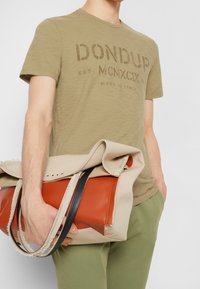 Beige och orange tygväska med en svart rem; har en strukturerad canvaskropp och en rullad topp. Man i olivgröna byxor håller väskan.