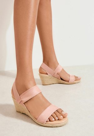 Sandały na koturnie z jasnoróżowymi paskami i plecionymi obcasami z liny, noszone na opalonych stopach, stojące na gładkiej jasnej podłodze.