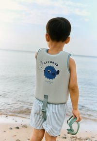 Svītrainas pelēkas un zilas bērnu peldvestes ar zivs grafiku. Bērns valkā svītrainas šortus, tur piekrastē zaļas peldbrilles.