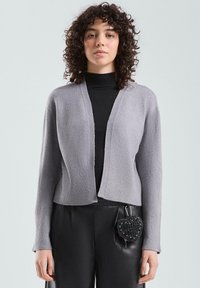 Szary krótki kardigan z dekoltem w serek, teksturowaną tkaniną i ściągaczami na rękawach, zestawiony z czarnymi spodniami i torebką w kształcie serca.