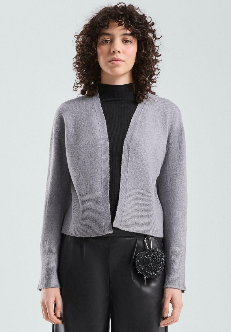Szary krótki kardigan z dekoltem w serek, teksturowaną tkaniną i ściągaczami na rękawach, zestawiony z czarnymi spodniami i torebką w kształcie serca.