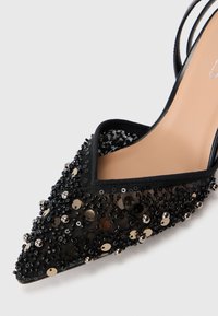 Musta kärjellä oleva korkokenkä, jossa on verkkohuppu, joka on koristeltu mustilla helmillä, paljeteilla ja metallisilla yksityiskohdilla, sekä vaaleanruskea pohja.