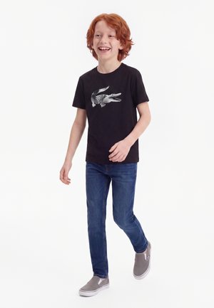 Garçon roux marchant en souriant, portant un t-shirt noir avec un motif de crocodile, un jean bleu et des chaussures grises sans lacets.