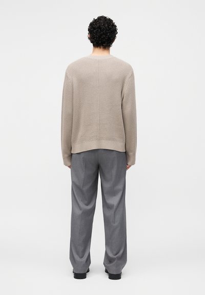 Homme aux cheveux bouclés portant un pull en maille beige, un pantalon gris à jambes larges et des chaussures noires, debout, dos tourné, sur un fond uni.