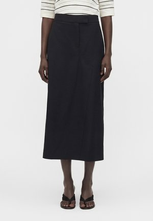 Γυναίκα φοράει μια μαύρη midi φούστα με κρυφό μπροστινό κλείσιμο, συνδυασμένη με κρεμ πλεκτό τοπ και μαύρα σανδάλια.