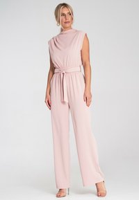Różowy kombinezon wykonany z gładkiego materiału, z wysokim kołnierzem, bez rękawów i elastycznym pasem z detalem w postaci wiązania; szerokie nogawki.