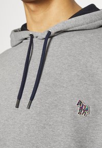 Sweat à capuche gris avec des cordons bleu foncé et un petit zèbre multicolore brodé sur la poitrine, porté par une personne montrée de la nuque jusqu'en bas.