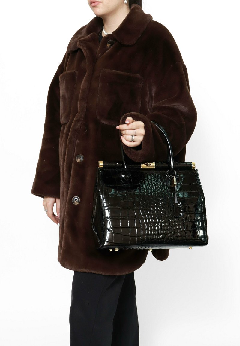 Borsa nera lucida con texture in coccodrillo e hardware dorato, tenuta da una persona che indossa un cappotto in pelliccia sintetica marrone con due tasche frontali.