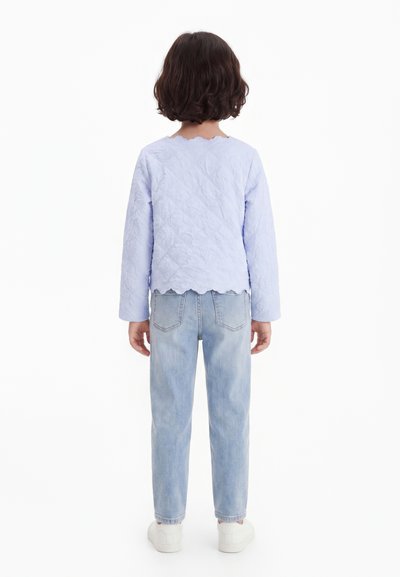 Copil cu păr scurt și închis la culoare, purtând un top texturat albastru deschis cu mâneci lungi, blugi albastru deschis și adidași albi, stând cu spatele.