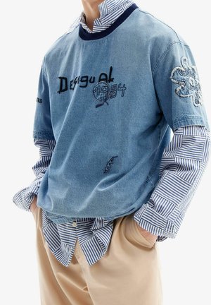 Personne portant une chemise à rayures bleues superposée sous un sweat-shirt bleu brodé à manches courtes et un pantalon beige, les mains dans les poches.