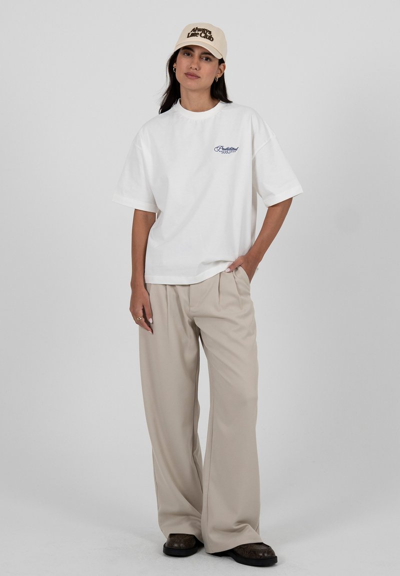 Femme debout avec une main dans la poche d'un pantalon beige à jambes larges, portant un t-shirt blanc surdimensionné et une casquette beige « Always Late Club ».