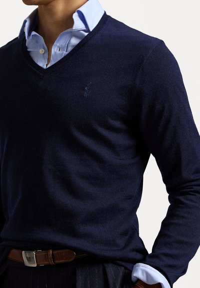 Suéter de punto azul marino con diseño de escote en V, que presenta un pequeño logo en el pecho. Lleva encima una camisa de color azul claro con cuello y un cinturón marrón.