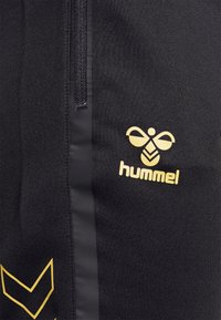 Tissu noir avec fermeture éclair verticale et logo hummel jaune avec une abeille stylisée et des chevrons sur le côté gauche.