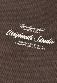 Καφέ ύφασμα με λευκό κείμενο που λέει Originals Roots Global Reach, Originals Studio, Everyday Essentials Creative Dept. Holmen.