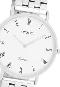 Montre bracelet en argent avec un cadran rond blanc, des chiffres romains et un bracelet en métal texturé. Présente le nom de la marque "OOZOO" et un détail "Vintage".