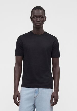 Mand med mørk hud iført en ensfarvet sort kortærmet t-shirt og lyseblå jeans, stående mod en ensartet lys baggrund.