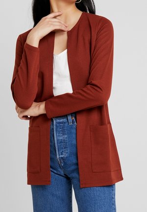 Vrouw draagt een roestkleurige cardigan met open voorkant en zakken over een wit shirt en blauwe spijkerbroek, hand raakt haar nek aan en arm is gekruist.