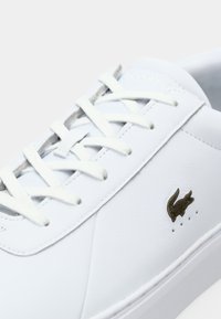 Sapatilhas brancas de atacador com um acabamento em pele lisa, apresentando um logo de crocodilo em bronze e perfurações subtis na lateral.