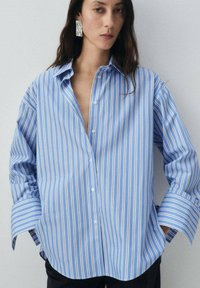 Camicia a righe blu e bianche con una vestibilità ampia, caratterizzata da maniche lunghe e colletto a punta. Tessuto di cotone liscio, senza fantasie.