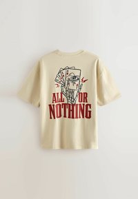 T-shirt en coton beige à manches courtes avec un graphisme d'une main squelettique tenant des cartes à jouer, avec le texte rouge "TOUT OU RIEN."