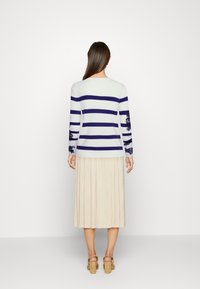 White sweater with navy stripes and floral lace accents on sleeves, paired with a pleated beige skirt, and tan block-heeled sandals.