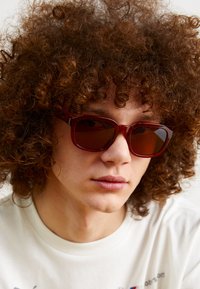 Gafas de sol rojas y rectangulares con lentes oscuros. Montura de plástico grueso, textura suave y bordes redondeados. Llevadas por una persona con cabello rizado.
