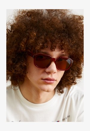Lunettes de soleil rouge, rectangulaires avec des verres foncés. Monture en plastique épais, texture lisse et bords arrondis. Portées par une personne avec des cheveux bouclés.