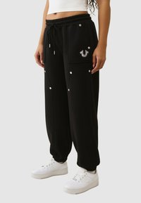 Zwarte joggerbroek gemaakt van zacht materiaal, met meerdere zakken, zilverkleurige accenten en een decoratief logoplaatje.