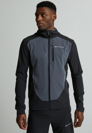 Homme portant une veste zippée Monterrain noire et grise avec capuche et un pantalon noir assorti, regardant vers sa gauche.