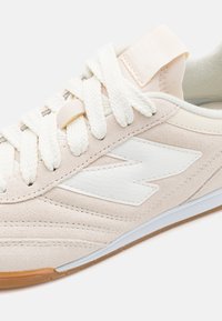Primer plano de una zapatilla de ante beige con un logo de cuero blanco, cordones blancos gruesos y borde de suela de goma marrón claro.
