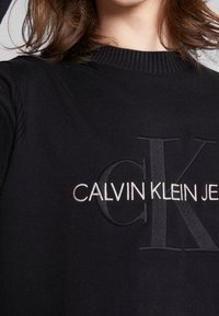 Černý pletený svetr s ribbovaným výstřihem. Vyšívaný nápis "CALVIN KLEIN JEANS" v kontrastních krémových a šedých tónech.
