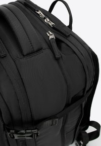 Zaino nero con struttura imbottita, scomparti multipli con zip, spallacci regolabili e tessuto resistente. Presenta una fibbia laterale e una texture liscia.