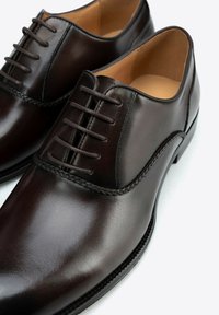 Los zapatos de vestir de piel marrón presentan un acabado suave, un sistema de cordones cerrado, acentos cosidos y un diseño de puntera redondeada con una suela texturizada.