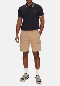 Polo di colore blu scuro con bordi bianchi, pantaloni cargo beige e scarpe da ginnastica grigie con accenti neri e bianchi. Il modello è in piedi contro uno sfondo semplice.