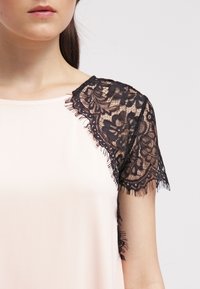 Blush blouse featuring short sleeves of black lace with floral patterns, fringe detailing along the edges, and a smooth fabric body.