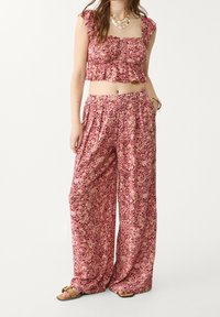 Vrouw in een roze patterned ruffled crop top en bijpassende wijde broek, accessorized met een gelaagde ketting en sandalen.