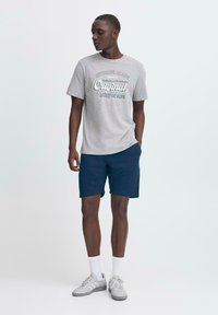 Camiseta de algodón gris con un estampado gráfico, combinada con pantalones cortos navy. El modelo lleva calcetines blancos y zapatillas grises, mostrando un look casual.