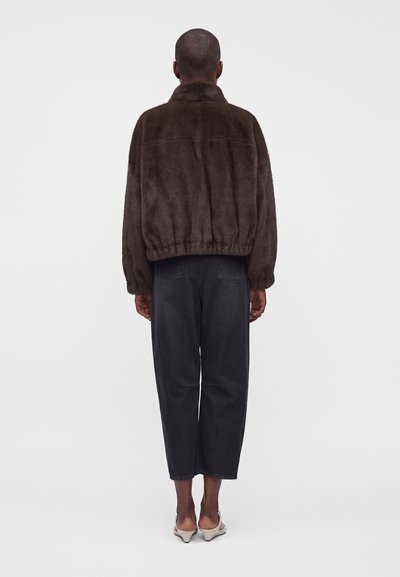 Personne de dos portant une veste en fourrure marron foncé, un jean noir ample et des chaussures à talons claires sur fond blanc.