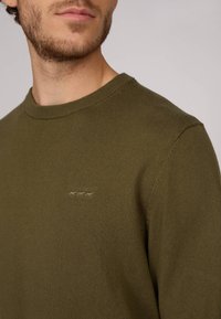 Pull en vert olive fabriqué en tissu tricoté, avec un col rond et un logo brodé discret sur la poitrine. Texture lisse, design ajusté.