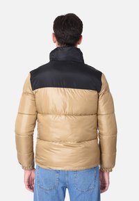 Giacca puffa di colore beige con yoke nero, texture trapuntata, colletto alto e polsini elasticizzati. Adatta per il tempo freddo.
