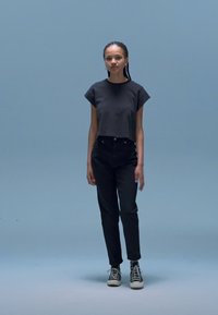 T-shirt preta cropped com mangas curtas, combinada com jeans pretos de cintura alta. Modelo usa ténis pretos. O fundo é azul claro.