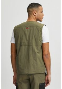 Vesta fără mâneci de culoare verde măslin, cu un finisaj texturat, având un inel roșu atârnat pe spate și un design simplu. Purtați peste o cămașă albă.